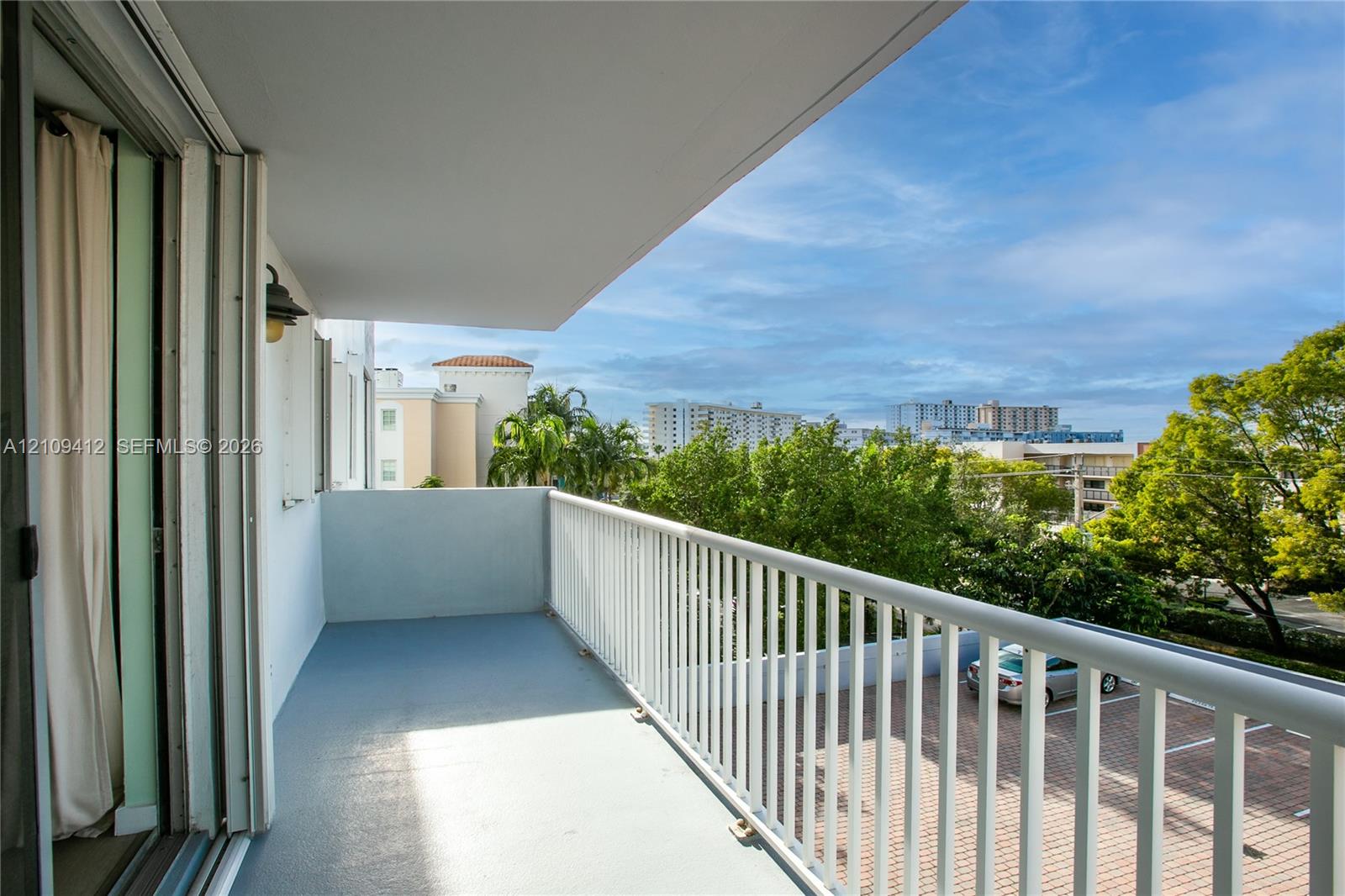 Covered outdoor balcony featuring a solid surface floor, white metal railing, and wall-mounted exterior light fixture