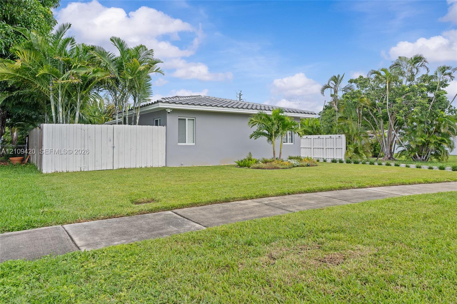 Single-story exterior with light gray siding, concrete tile roof, white privacy fencing, and established palm landscaping