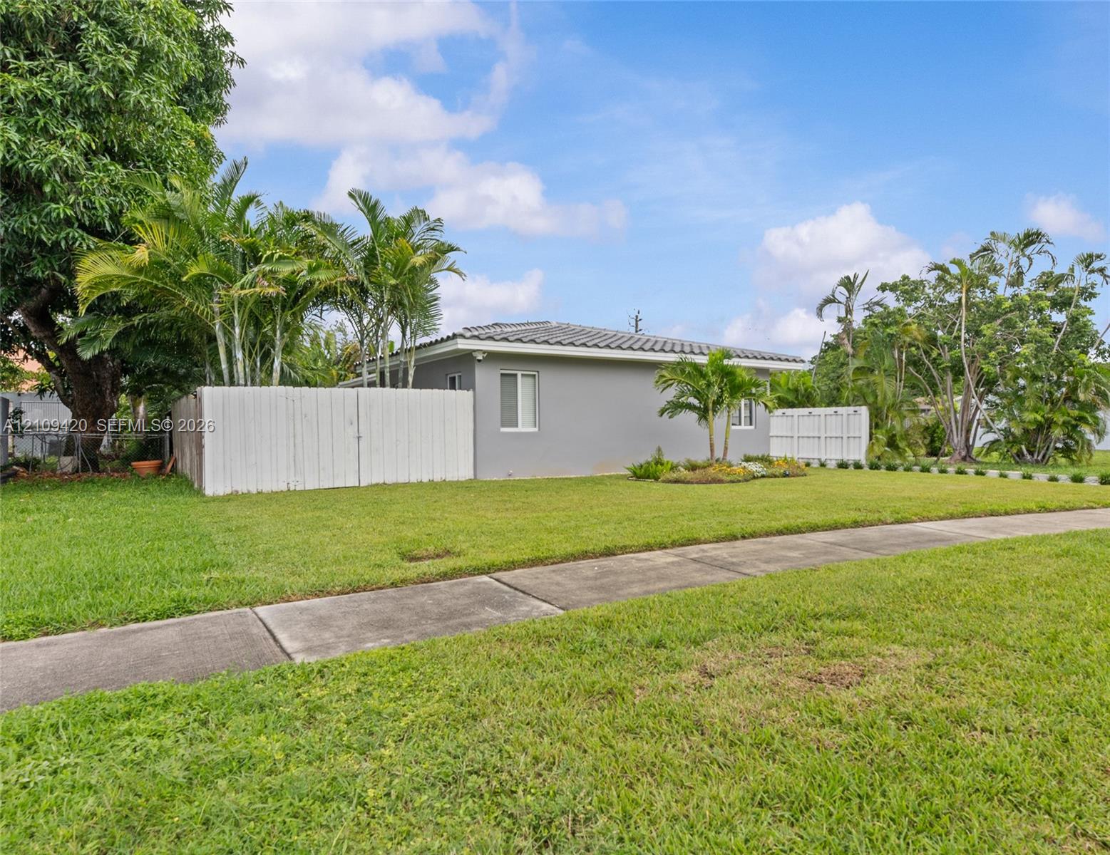 Exterior featuring a stucco facade, tile roofing, concrete sidewalk, and white fencing, accented by mature palm trees