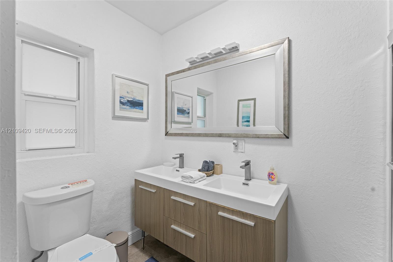 Dual-sink vanity featuring a wood-finish cabinet, rectangular basin sinks, brushed metal faucets, and a framed wall mirror with integrated lighting