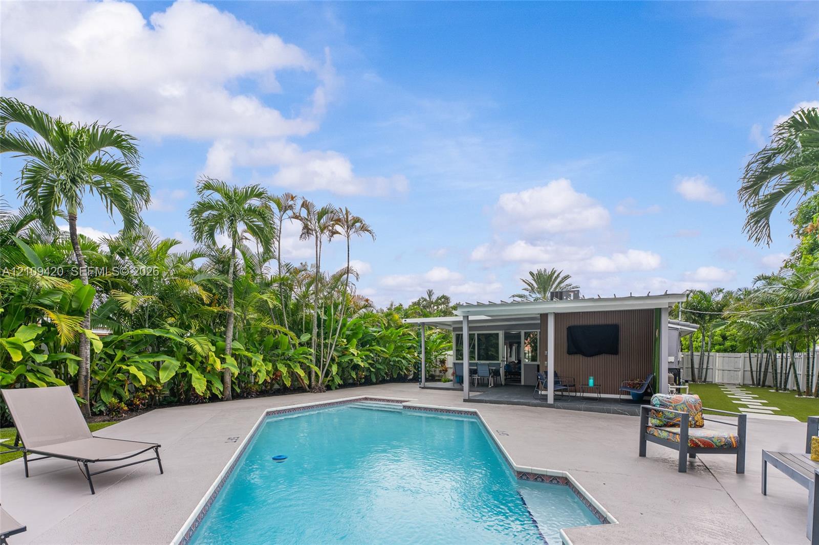 In-ground swimming pool featuring a shallow sun shelf, expansive concrete deck, covered patio with integrated pergola, and privacy-focused perimeter landscaping