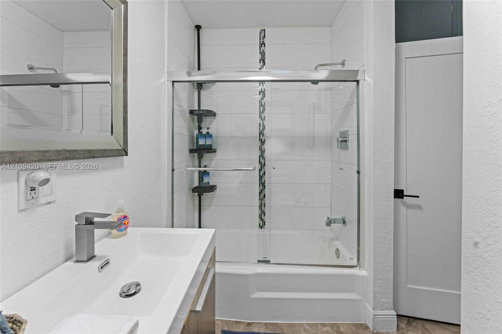 Modern vanity with a rectangular sink, wall-mounted mirror, glass-enclosed shower and tub combination featuring horizontal tile surrounds and a decorative mosaic accent strip