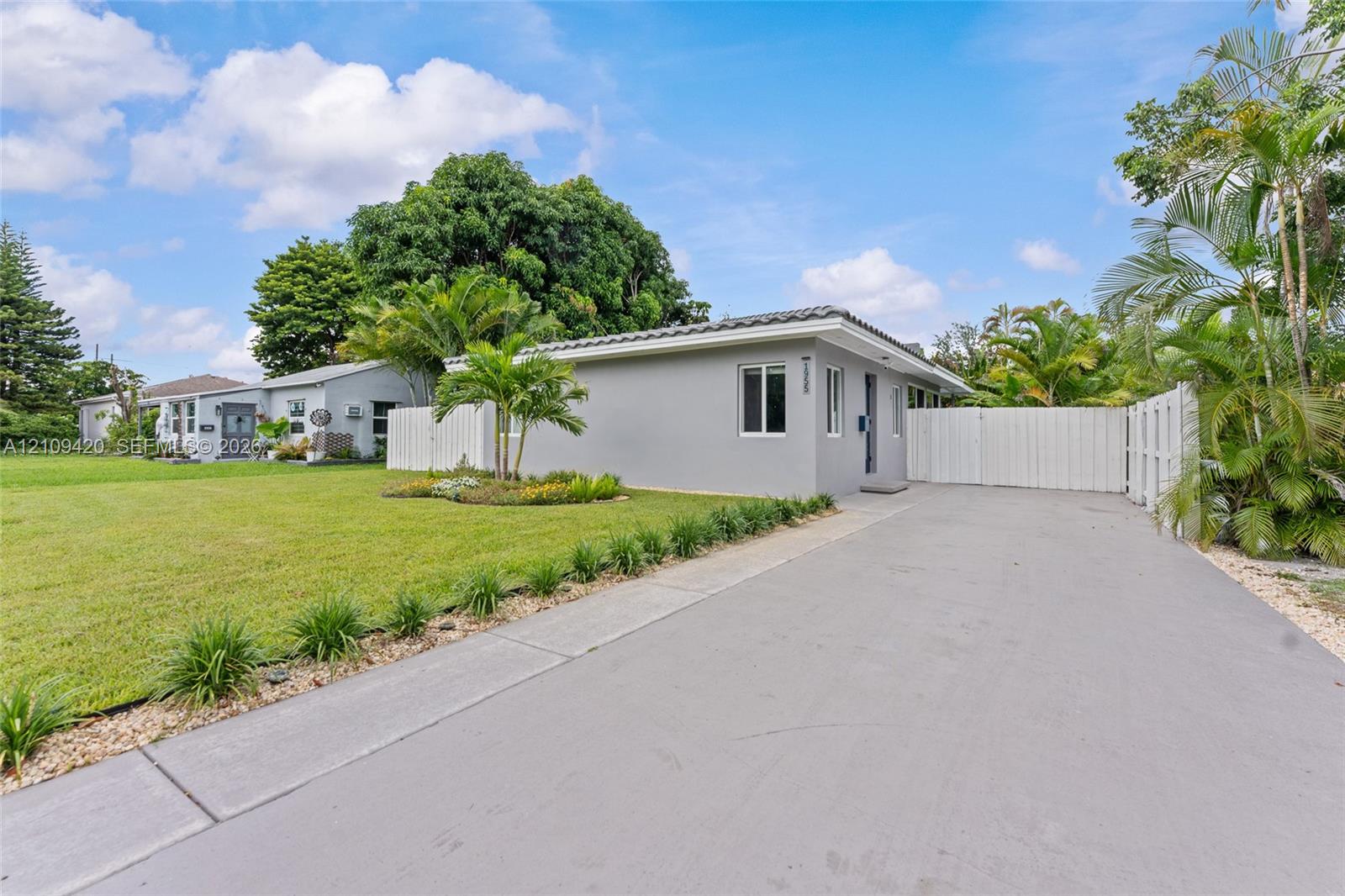 Single-story exterior featuring a concrete driveway, gated side entrance, manicured lawn, and established tropical landscaping