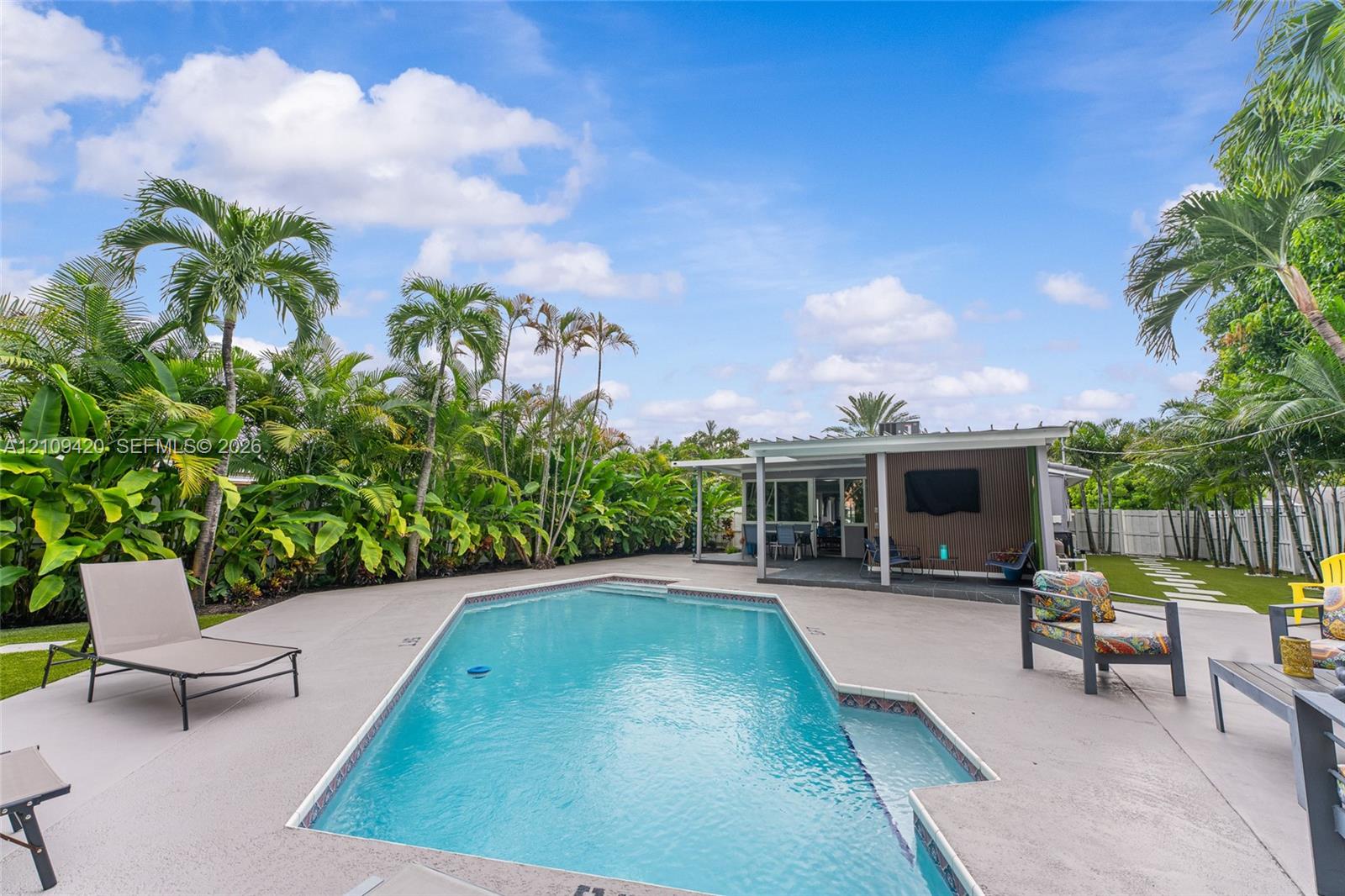 Backyard featuring an in-ground swimming pool, concrete pool deck, covered patio with structural support columns, and mature tropical landscaping