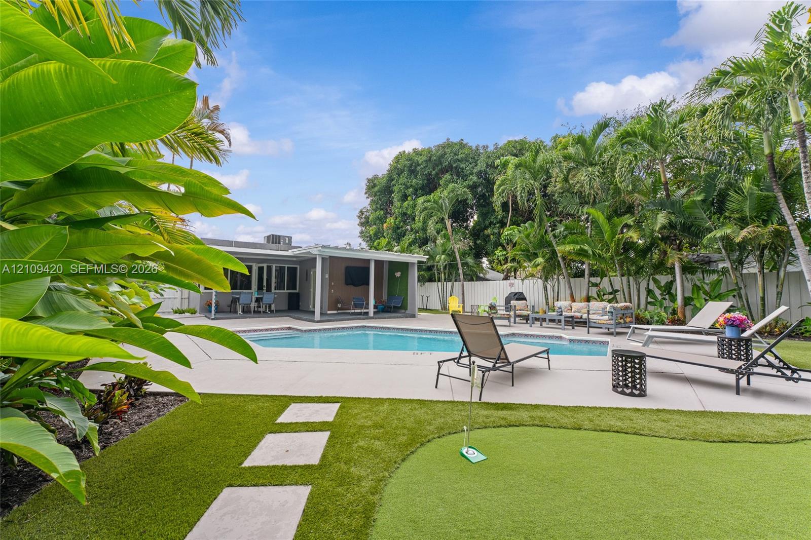 Backyard featuring an in-ground swimming pool, concrete pool deck, synthetic turf putting green, and mature tropical landscaping