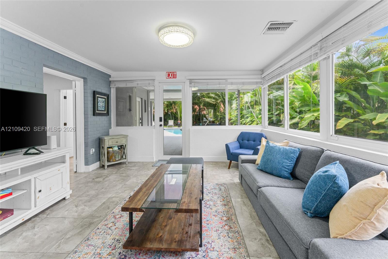 Sunroom featuring large-format tile flooring, blue-painted brick wall, flush-mount circular lighting, and extensive window enclosures with natural exterior scenery