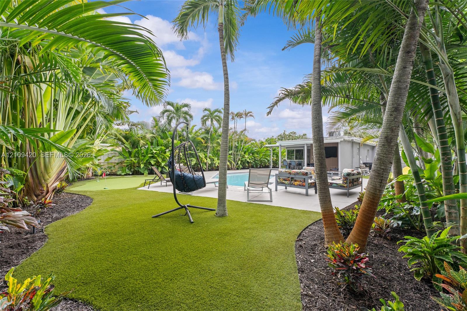 Backyard featuring a swimming pool, concrete patio, synthetic turf lawn, putting green, and mature palm trees