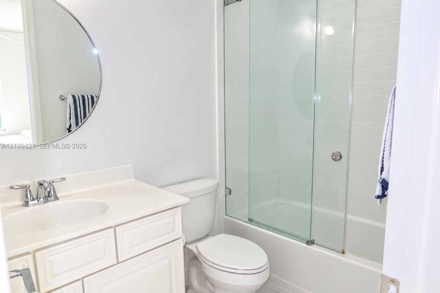 Bathroom featuring a vanity with an integrated sink and chrome faucet, circular wall mirror, and a tub-shower combination enclosed by sliding glass doors with tiled surround