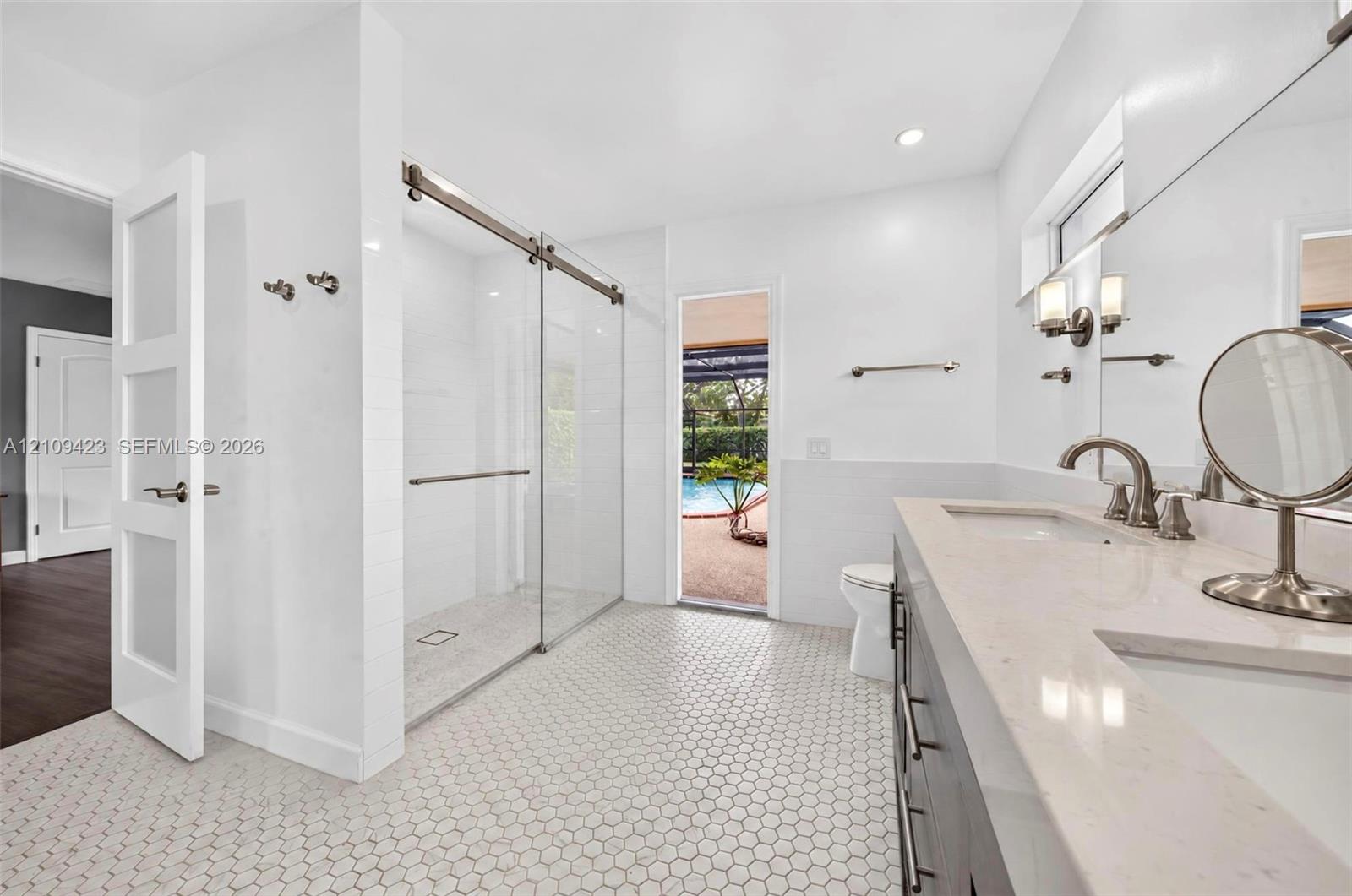 Dual-sink vanity with a solid-surface countertop, glass-enclosed walk-in shower with matte metal hardware, and hexagonal tile flooring