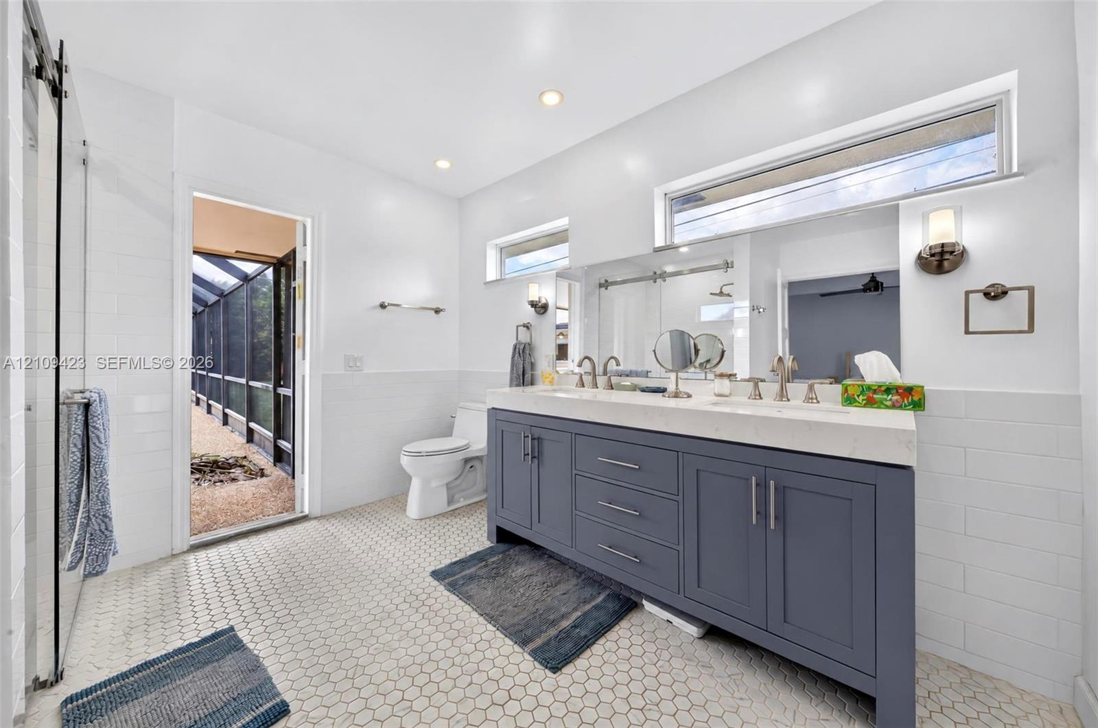 Dual-sink vanity with a quartz countertop, hexagonal tile flooring, glass-enclosed shower, wall-mounted light fixtures, and white subway-tile wainscoting