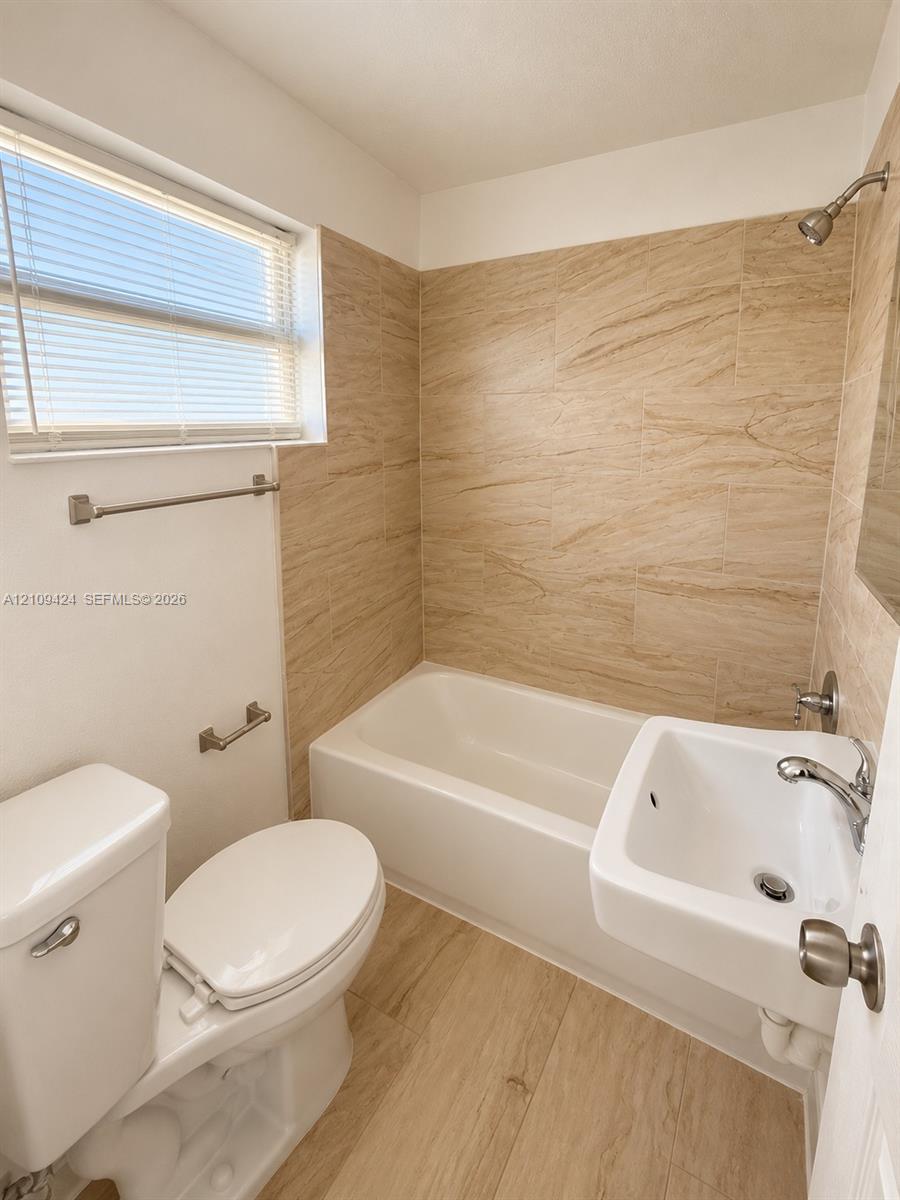 Bathroom featuring tile-surround bathtub, wall-mounted porcelain sink with chrome faucet, wood-finish flooring, and horizontal-slat window blinds