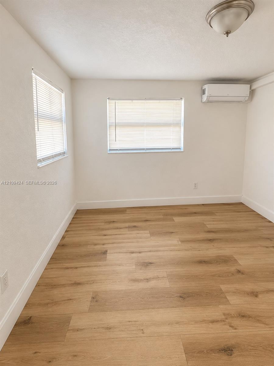 Neutral-toned room featuring wood-finish flooring, dual windows with horizontal blinds, and a wall-mounted ductless climate control unit