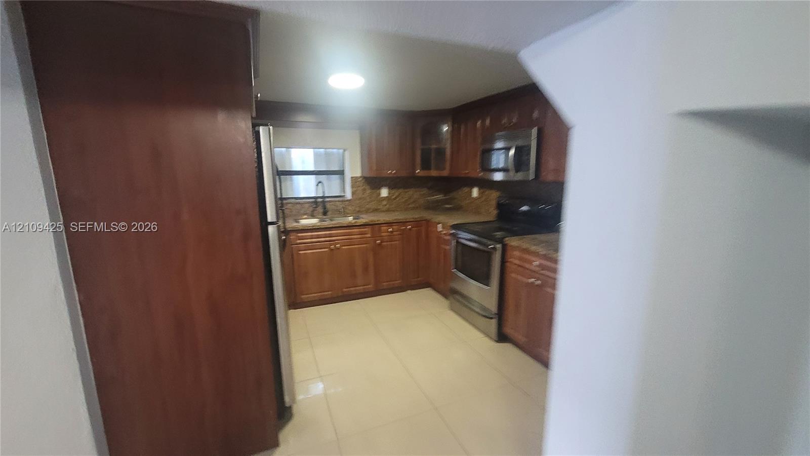 Kitchen featuring warm-tone wooden cabinetry, stone-finish countertops, stainless steel appliances, tiled backsplash, and light-tone tile flooring