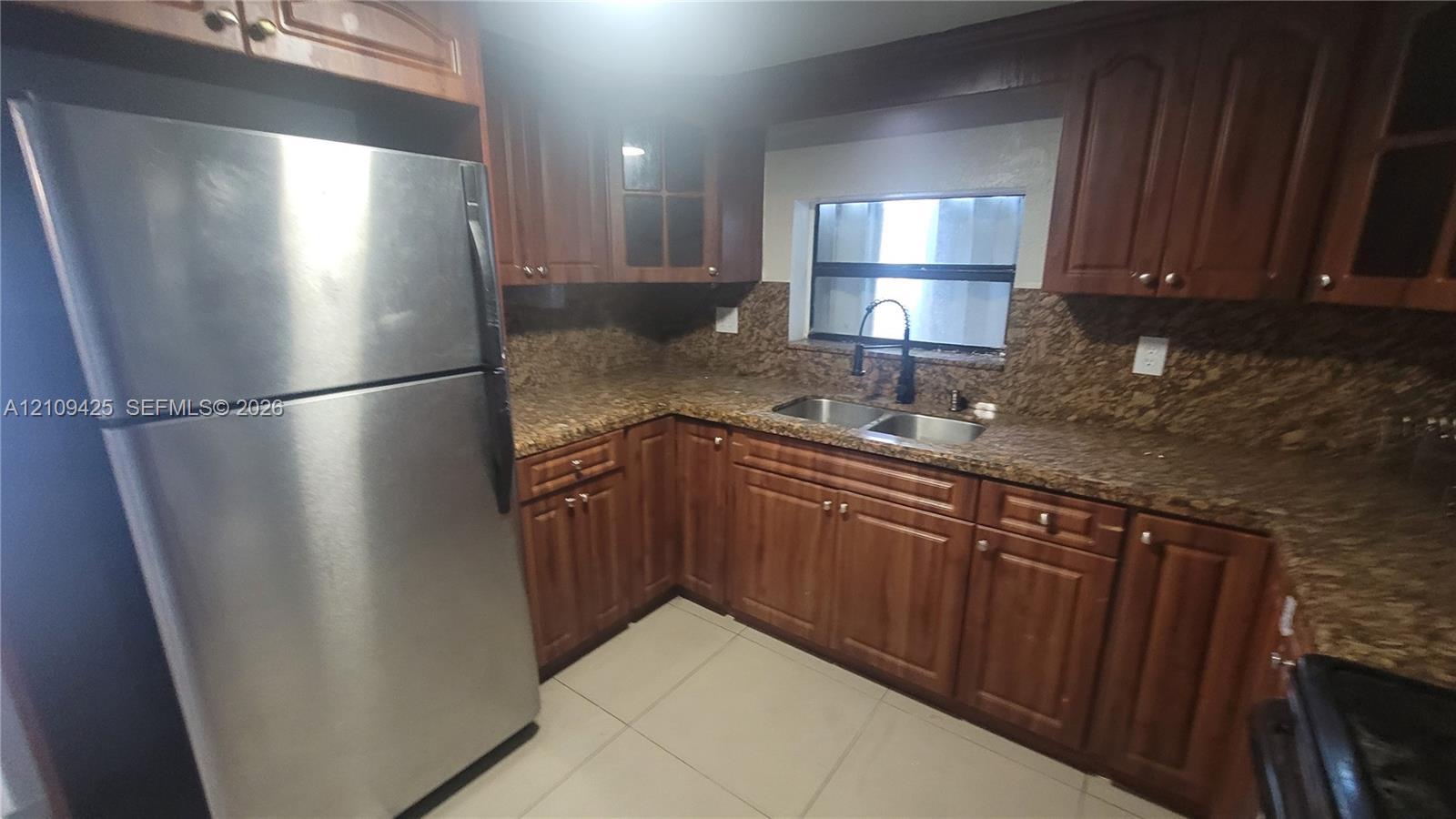 Kitchen featuring stained wood cabinetry, stone-surface countertops, dual-basin stainless steel sink with high-arc faucet, stainless steel refrigerator, and ceramic tile flooring