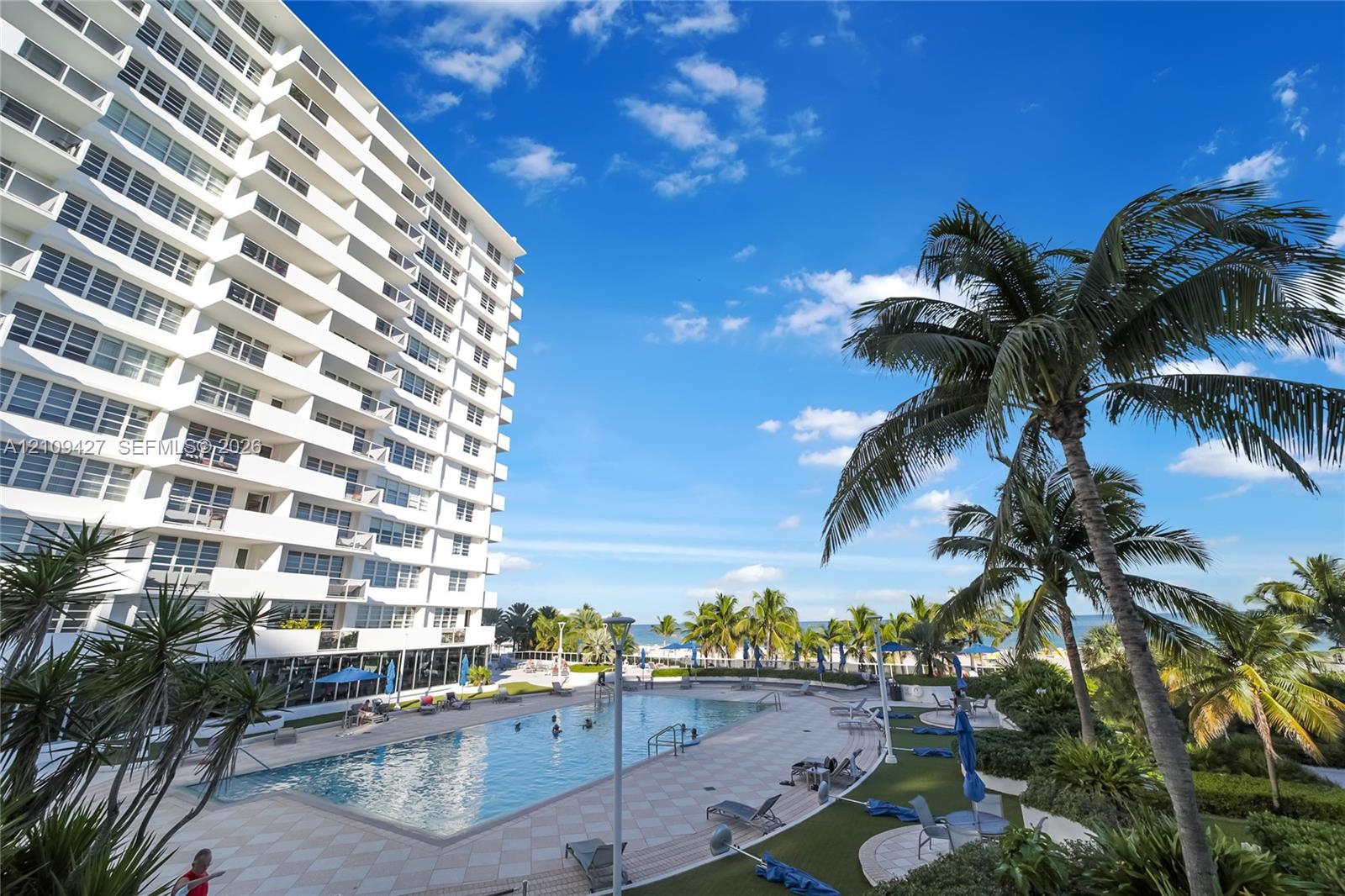 High-rise multi-story building featuring private balconies, an expansive ground-level swimming pool, and stone-paved decking surrounded by mature palm trees