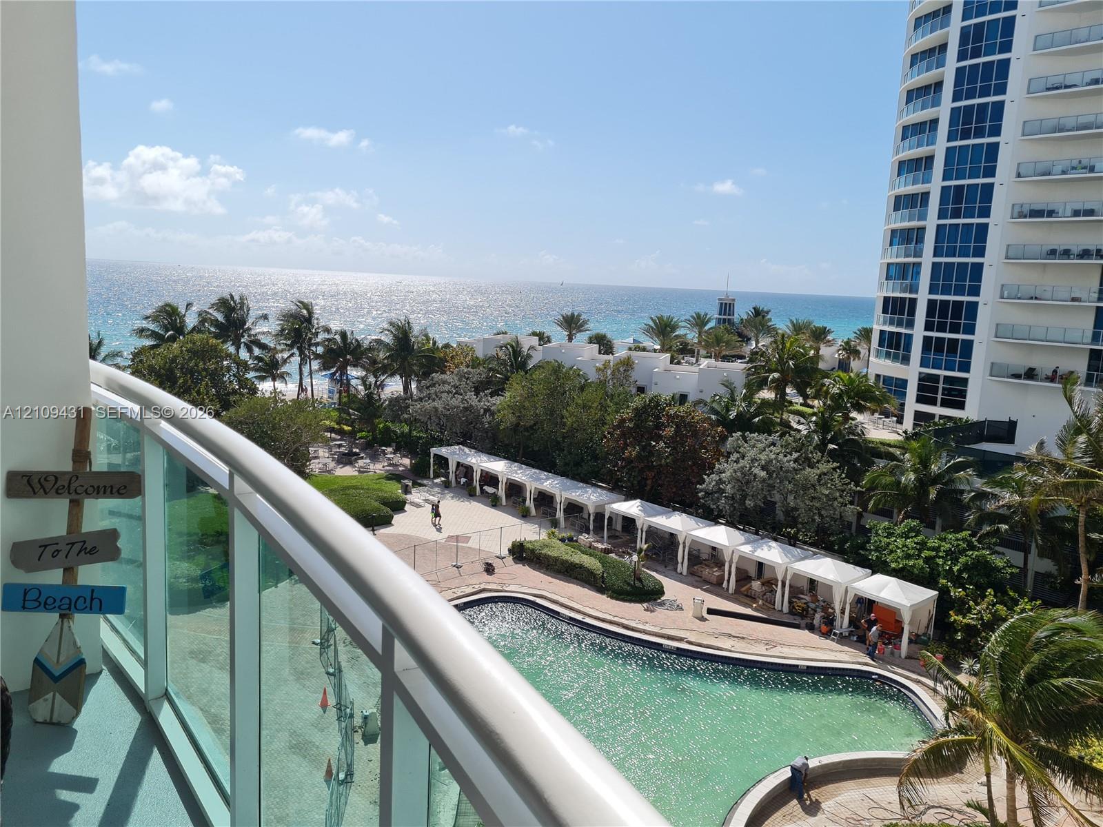 Balcony featuring a glass railing, overlooking an outdoor pool, cabanas, and ocean shoreline Balcony featuring a glass railing, overlooking an outdoor pool, cabanas, and ocean shoreline