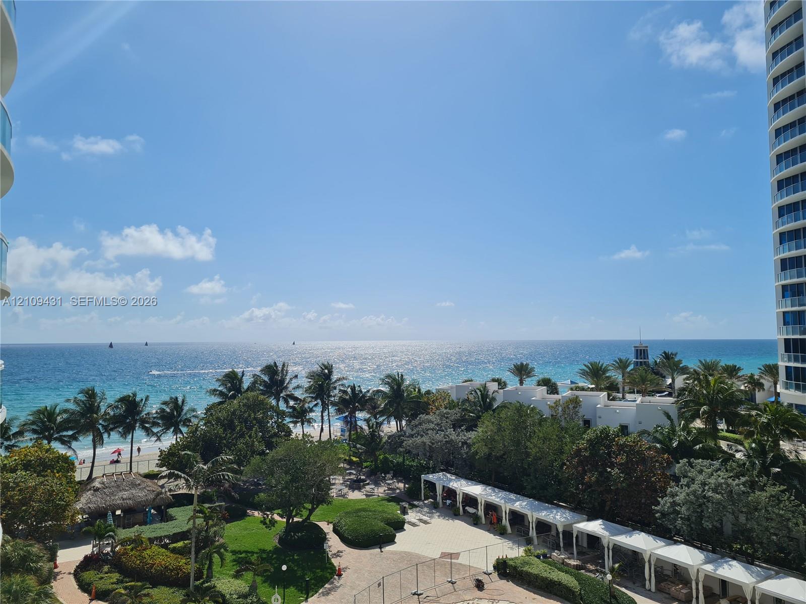 Oceanfront vista featuring mature palm trees, landscaped grounds, a paved courtyard, and covered white structural walkways