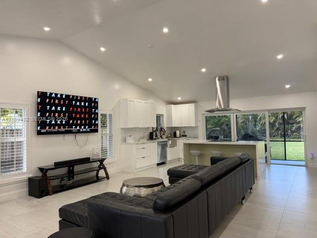 Open-concept kitchen and living area featuring high vaulted ceilings, recessed lighting, white shaker-style cabinetry, a central island with an integrated stainless steel range hood, and sliding glass doors