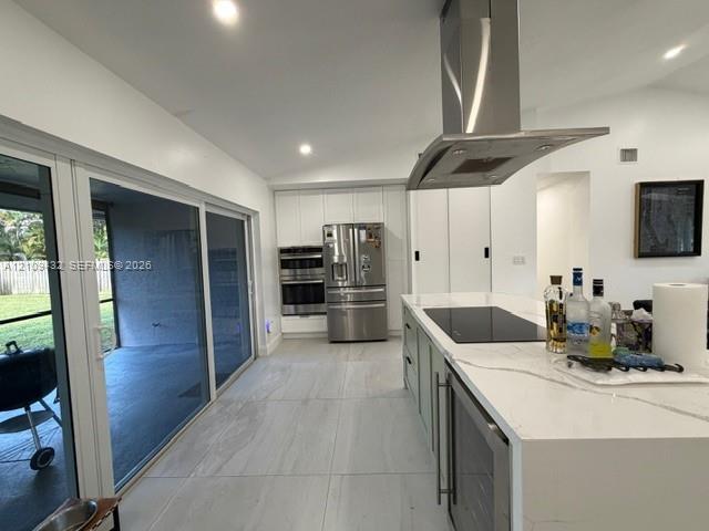 Kitchen featuring a large waterfall-edge island with an integrated cooktop and under-counter refrigeration, stainless steel wall-mounted range hood, custom cabinetry, stainless steel double wall ovens, and expansive sliding glass doors