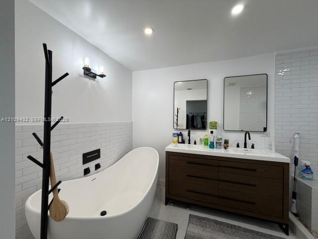 Freestanding soaking tub with wall-mounted matte black fixtures, double-sink vanity with a countertop, subway tile backsplash, and recessed lighting