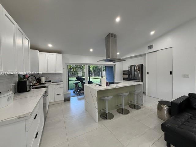Kitchen featuring a center island with a waterfall countertop, white shaker-style cabinetry, a stainless steel range hood, stone-finish flooring, and sliding glass doors