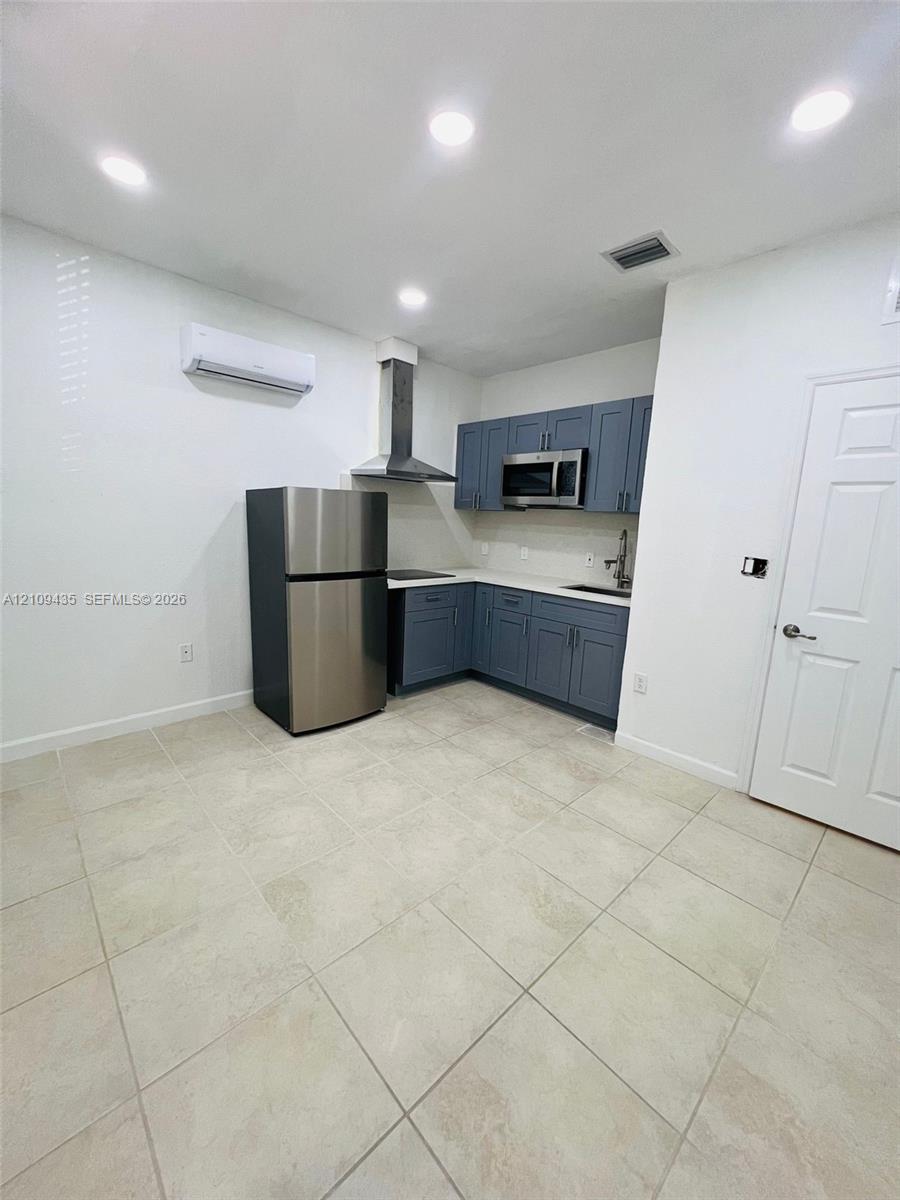Kitchen featuring blue shaker-style cabinetry, stainless steel appliances, a chimney-style range hood, solid-surface countertops, and light-tone tile flooring