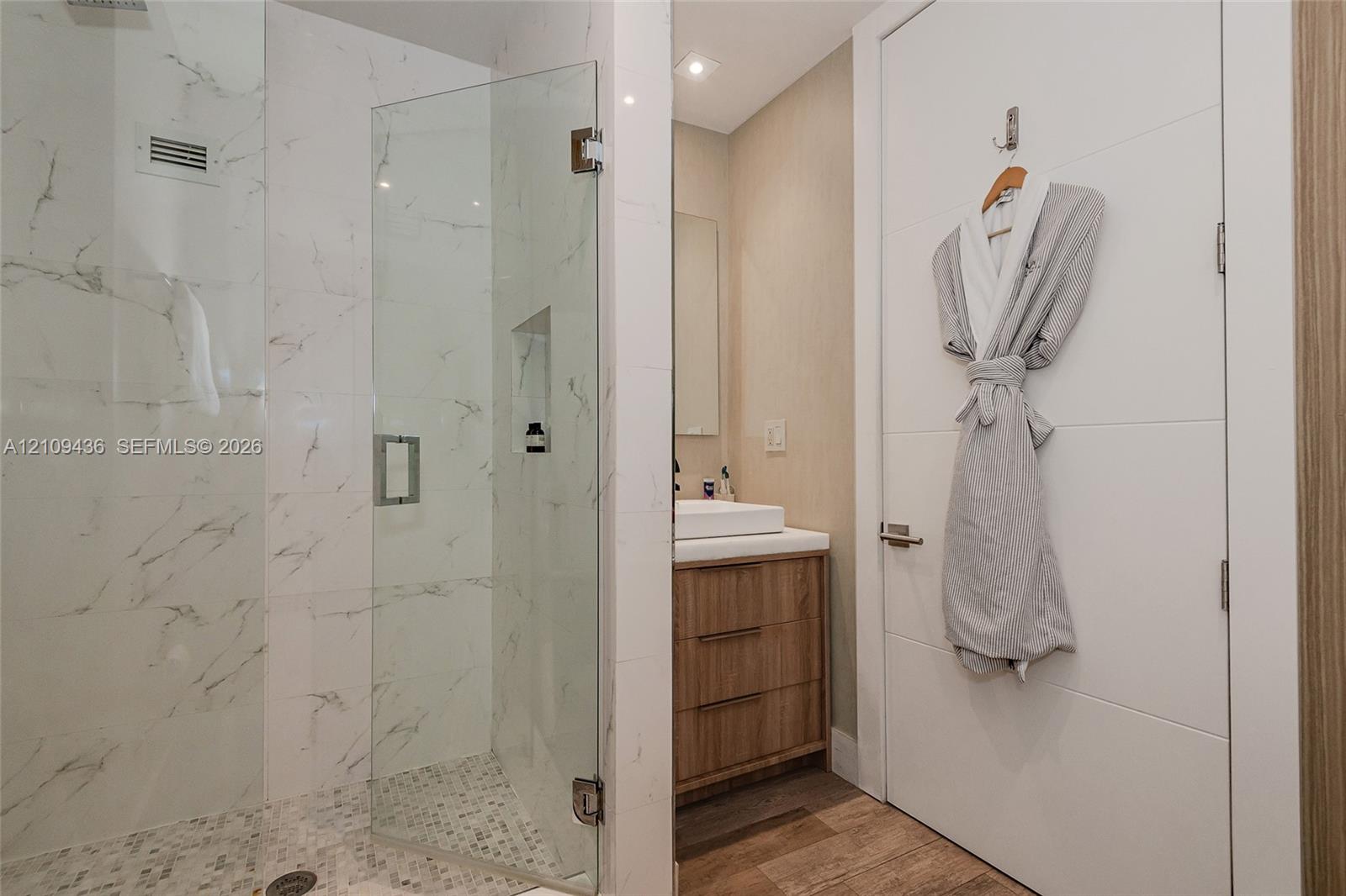 Walk-in shower featuring glass paneling, marble-style tile walls, and a pebble-stone floor, paired with a wood-finish vanity, vessel sink, and solid-core door