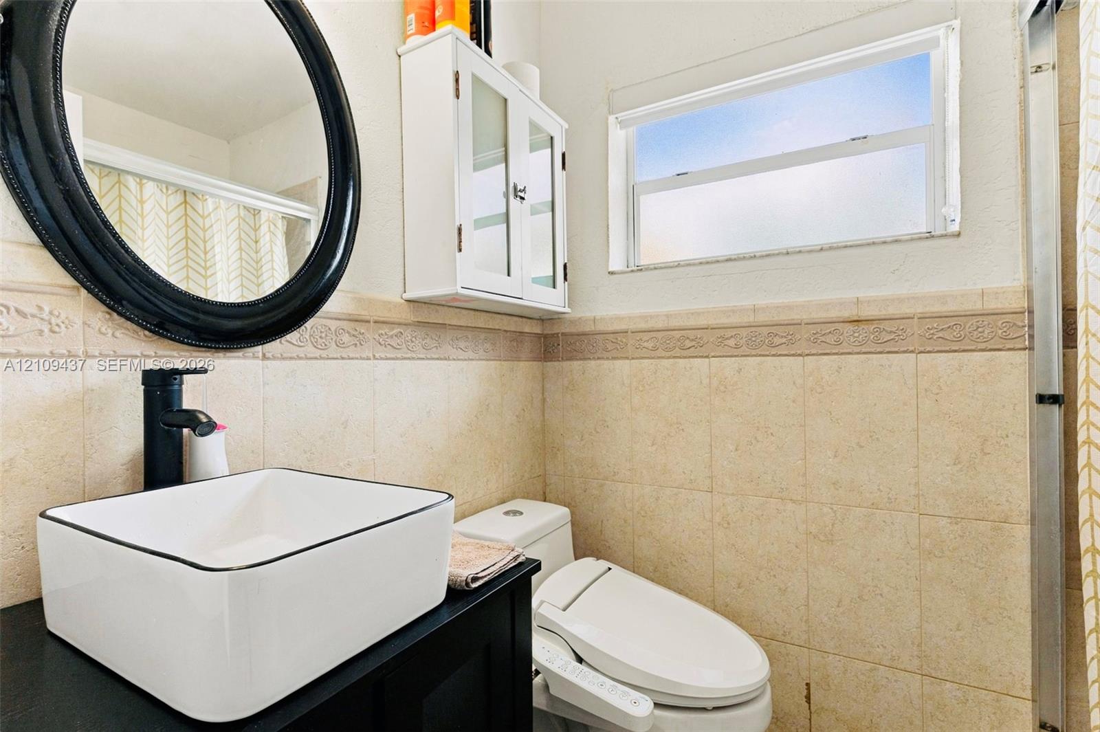 Rectangular vessel sink with matte black faucet, dark vanity cabinet, circular mirror, wall-mounted storage cabinetry, and tiled wall wainscoting