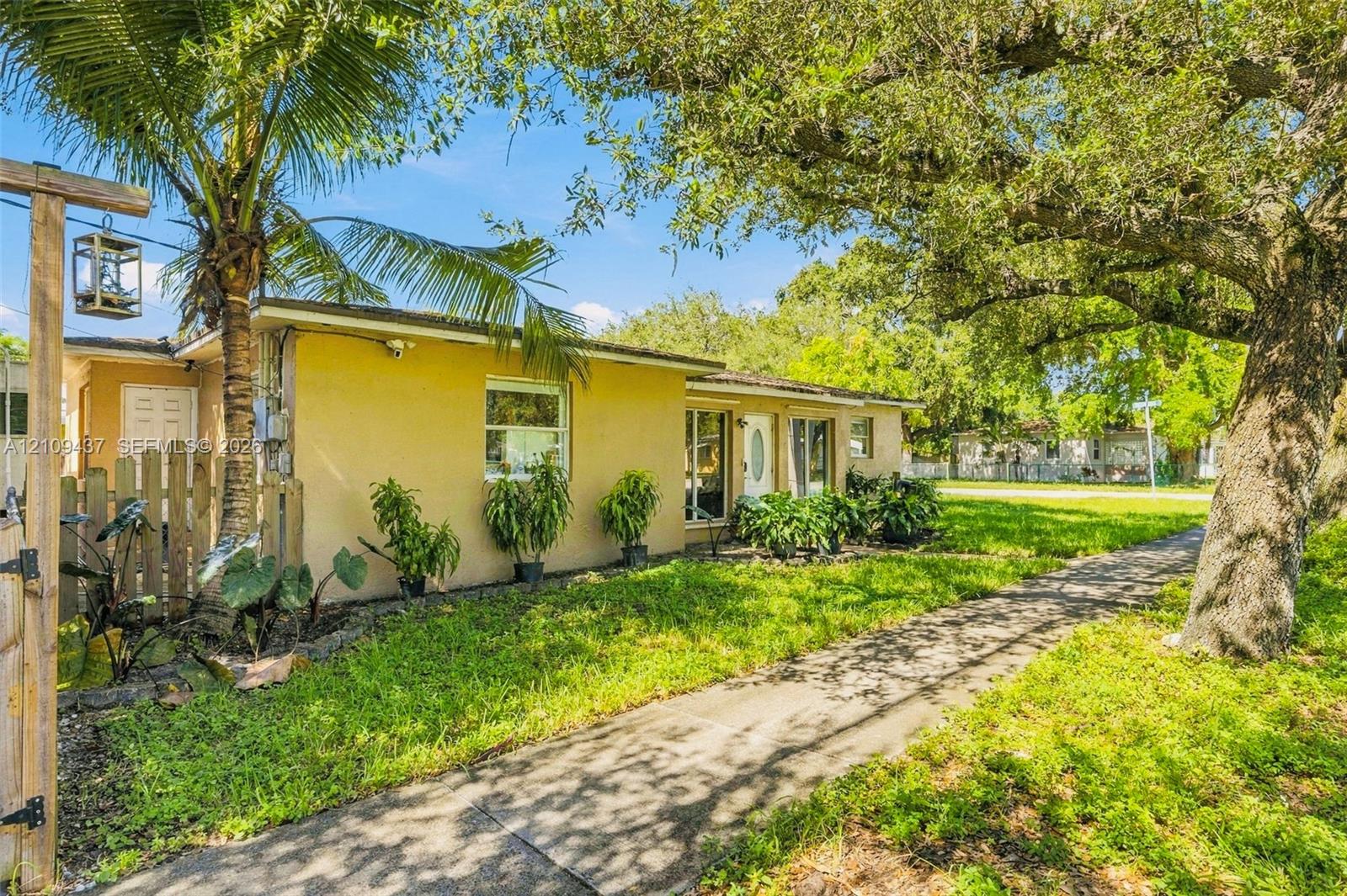 Single-story exterior featuring light-toned stucco siding, a concrete walkway, mature trees, and a wooden privacy fence Single-story exterior featuring light-toned stucco siding, a concrete walkway, mature trees, and a wooden privacy fence
