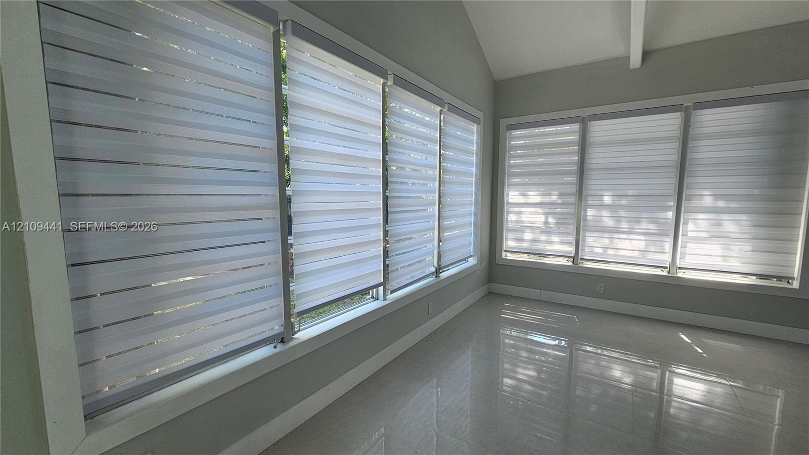 Vaulted ceiling featuring a central support beam, large windows with horizontal roller shades, and polished tile flooring