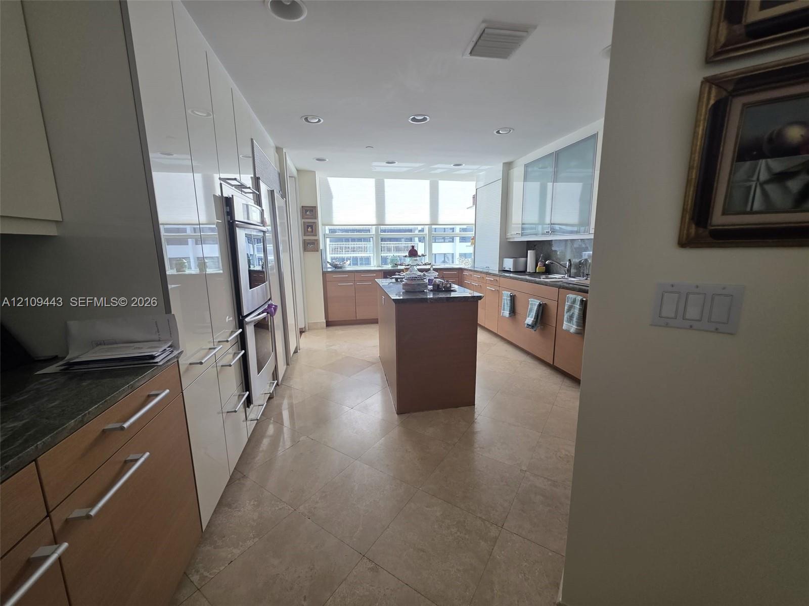 Kitchen featuring two-tone cabinetry with wood-finish lower units and high-gloss upper cabinetry, stone countertops, a central kitchen island, and large-format tile flooring