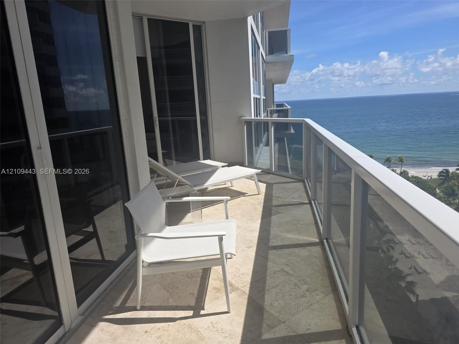 Covered balcony featuring stone tile flooring, floor-to-ceiling glass sliding doors, and a glass-paneled railing overlooking the ocean