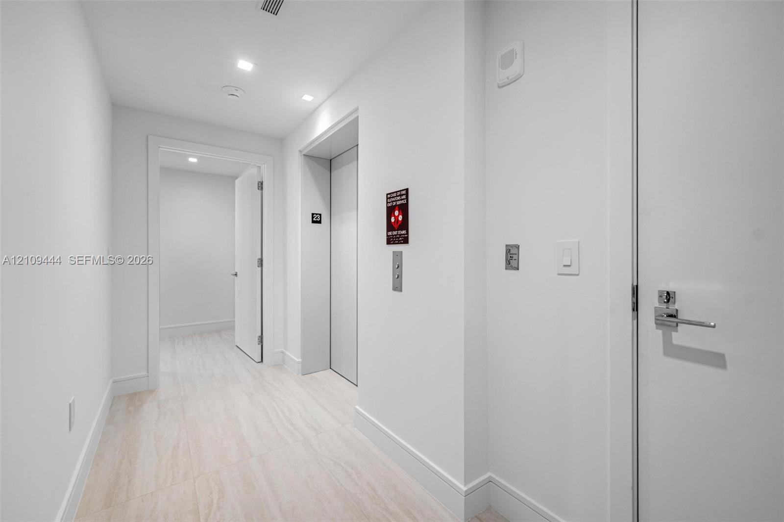 Neutral-toned interior hallway featuring recessed ceiling lighting, light-colored stone-finish flooring, elevator access, and solid-core interior doors with satin nickel hardware