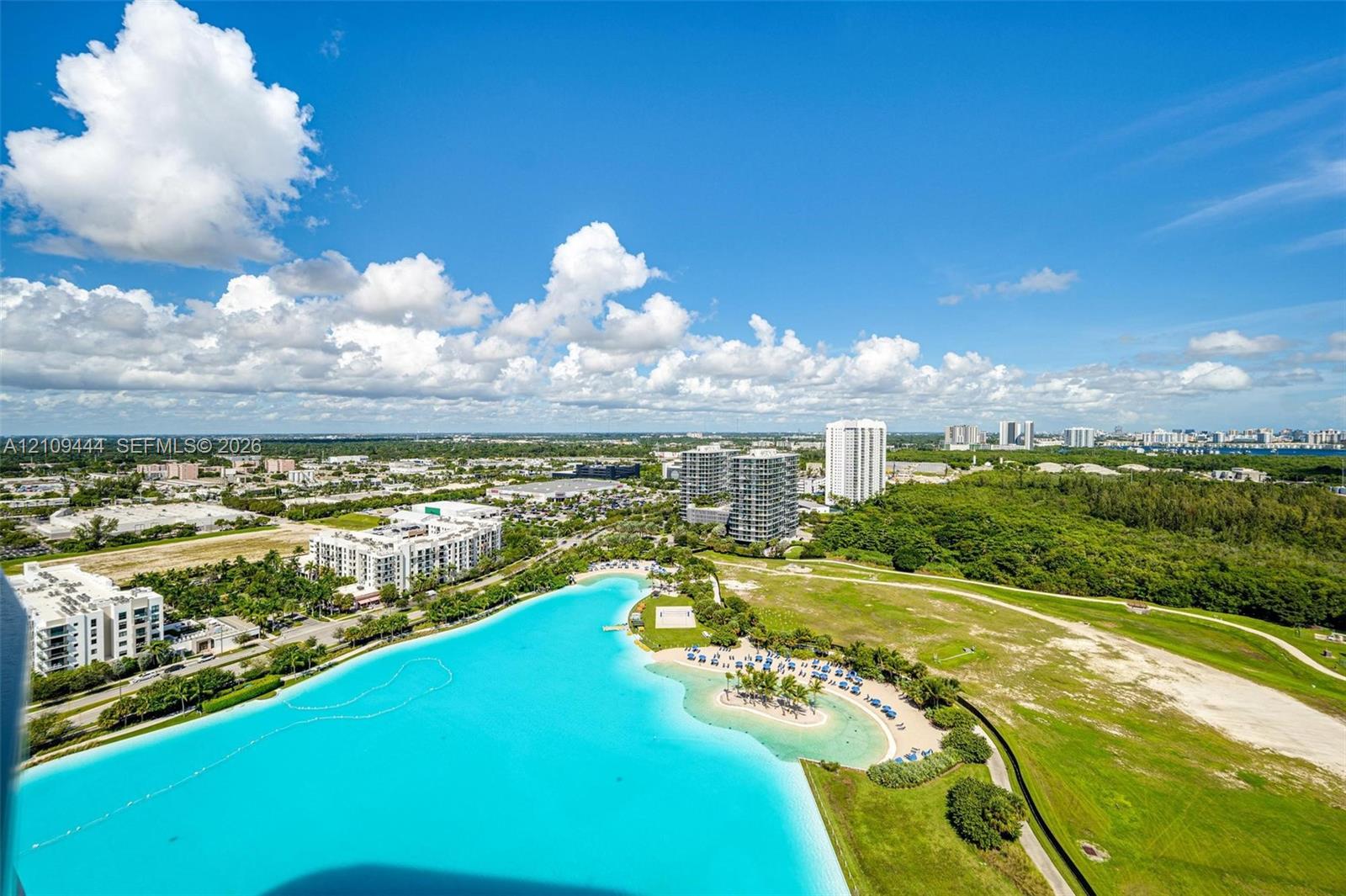 Aerial perspective featuring a large turquoise lagoon, sandy beach area, palm trees, and expansive green space