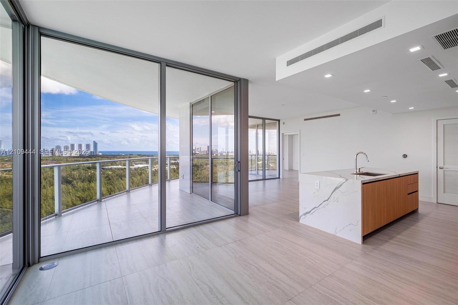 Floor-to-ceiling glass walls, expansive wraparound balcony, kitchen island with waterfall-edge stone countertop, wood-finish cabinetry, and neutral large-format tile flooring