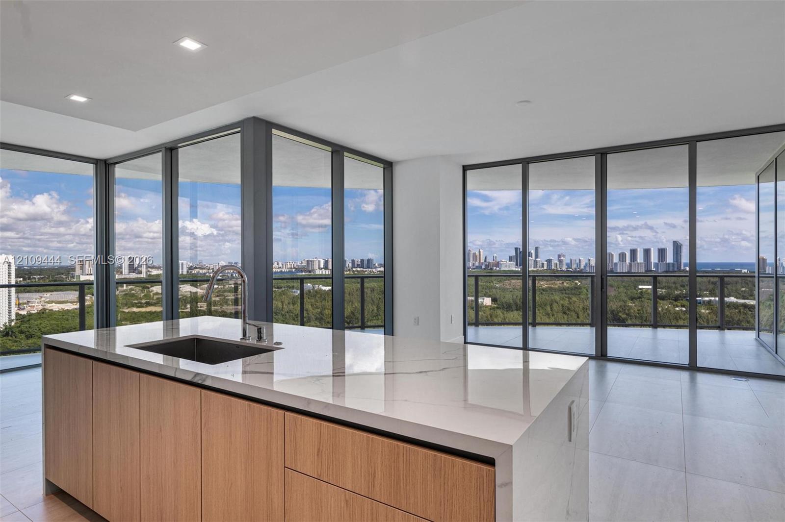 Kitchen island featuring waterfall-edge stone countertops, undermount sink, gooseneck faucet, and wood-finish cabinetry, set against floor-to-ceiling glass windows with expansive horizon scenery