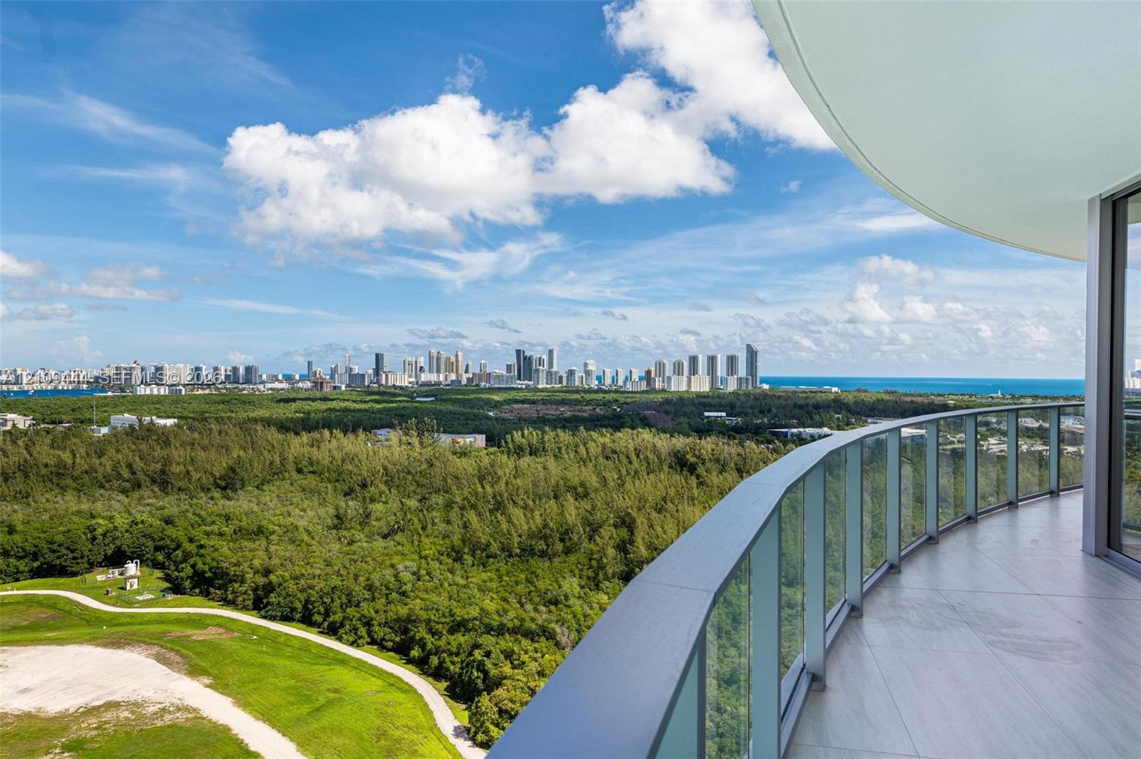 Curved exterior balcony with glass panel railing and tile flooring, featuring expansive sights of the coastal skyline and surrounding greenery