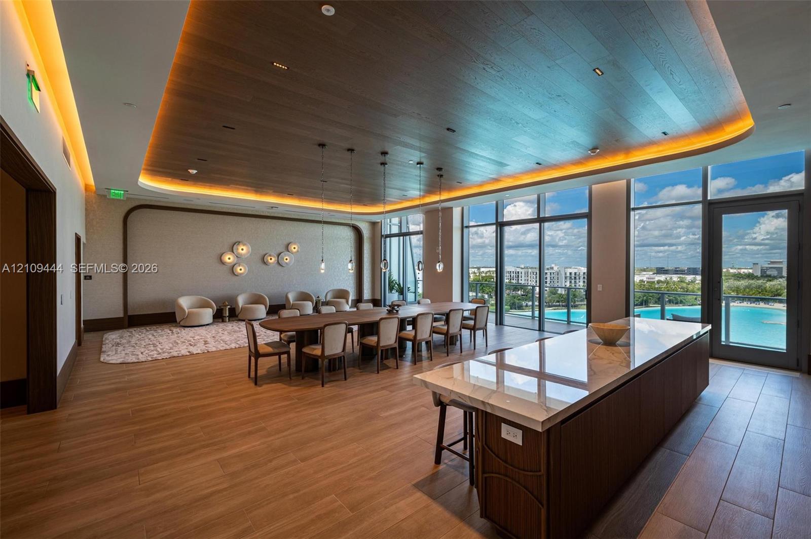 Expansive interior featuring a wood-paneled tray ceiling with recessed ambient lighting, floor-to-ceiling windows, wood-finish flooring, and a large kitchen island with a stone-finish countertop