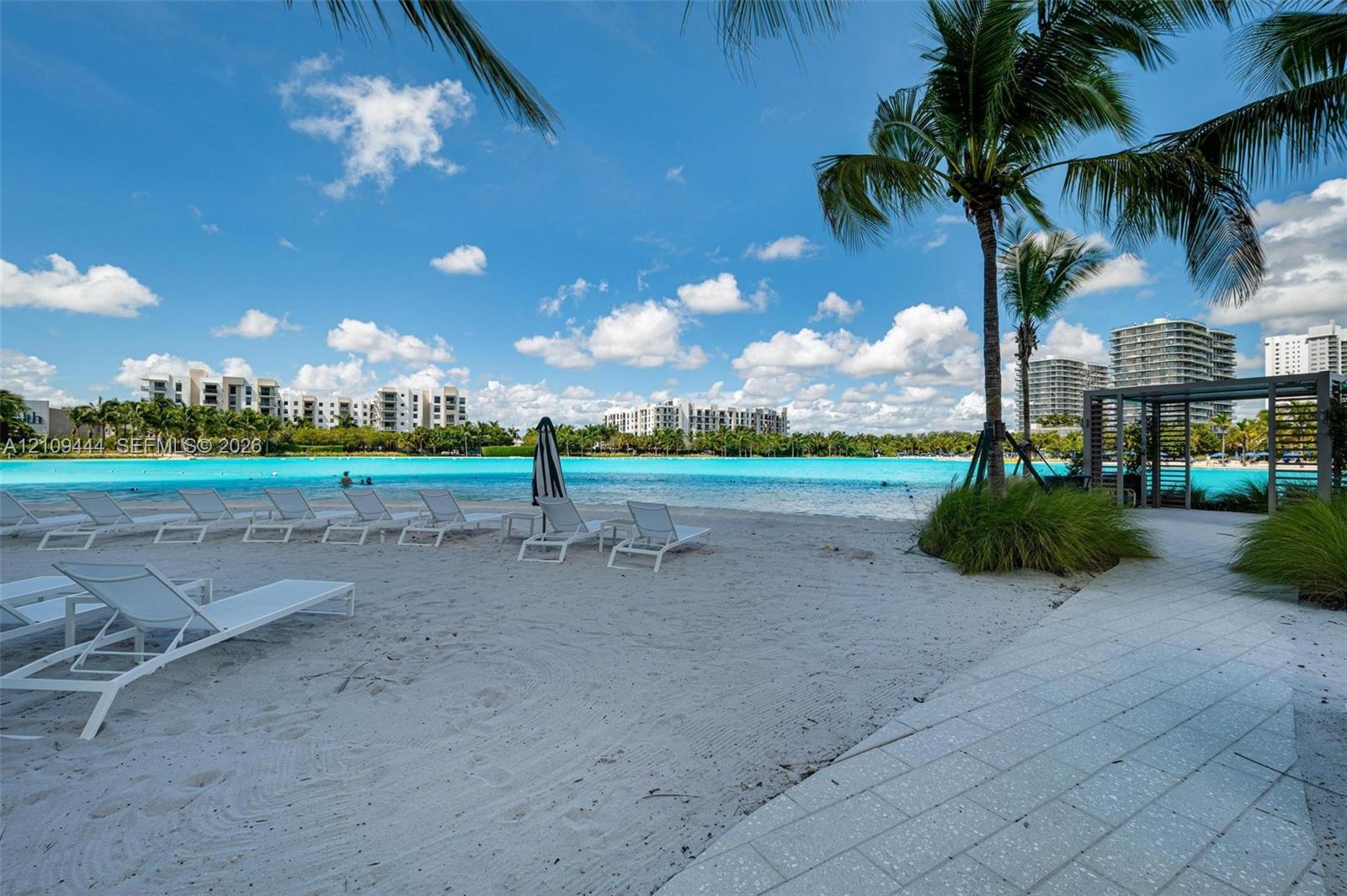 Sandy beach frontage featuring palm trees, a paved stone pathway, and an open-air pergola structure