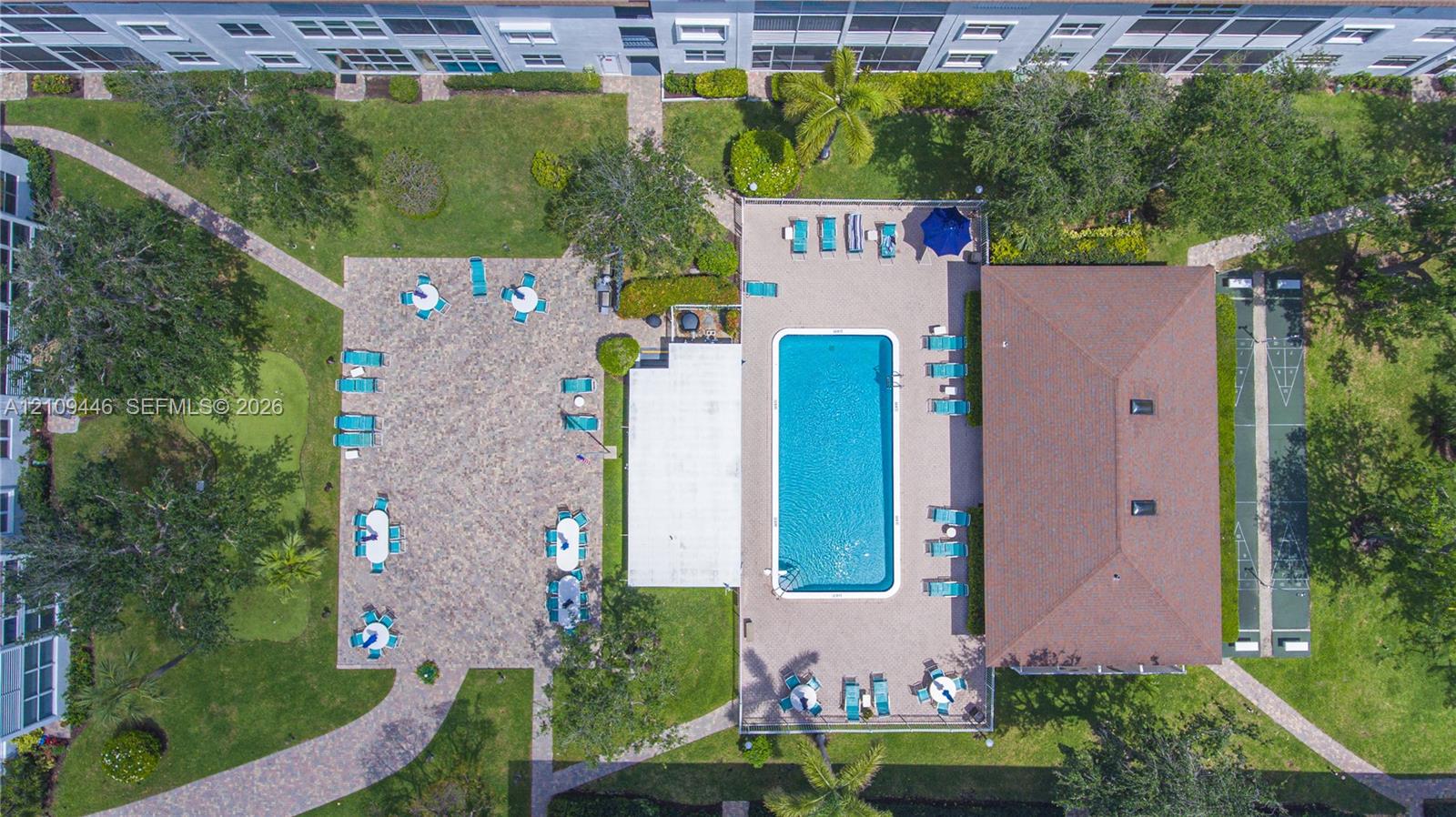Outdoor recreation area featuring a rectangular swimming pool, paver deck, shuffleboard court, and manicured green space with mature landscaping
