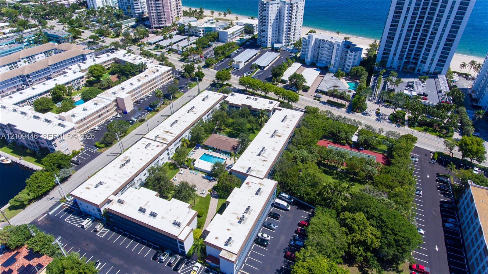 Aerial perspective of a multi-building residential complex featuring a central swimming pool, paved surface parking, mature landscaping, and a tennis court
