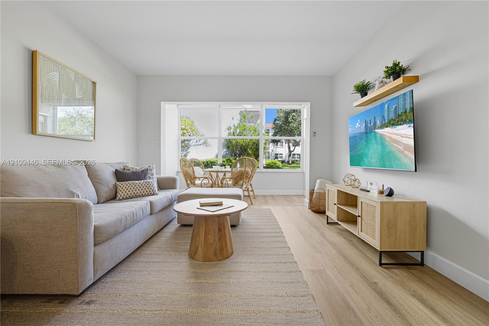 Living area featuring wood-finish flooring, neutral walls, large picture window with exterior greenery, and a wall-mounted floating shelf
