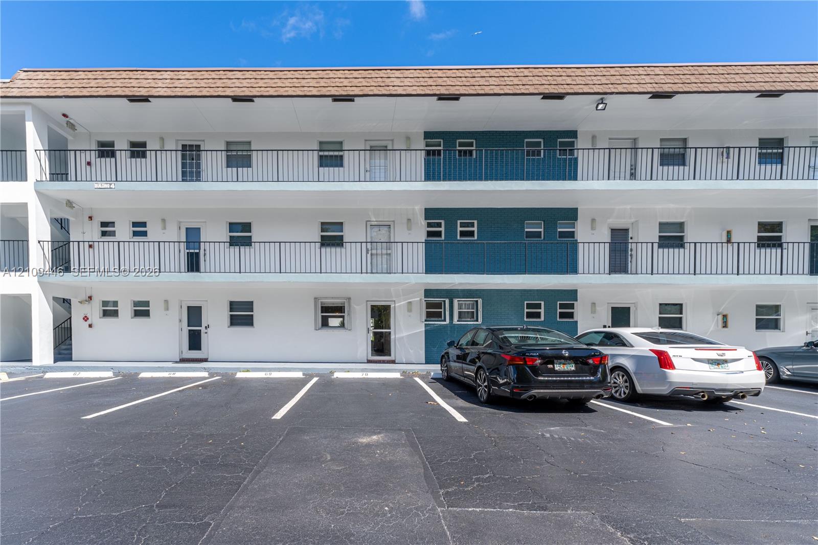 Multi-story exterior featuring white stucco siding, blue tile accents, metal balcony railings, and a tiled roof, complemented by a paved parking area
