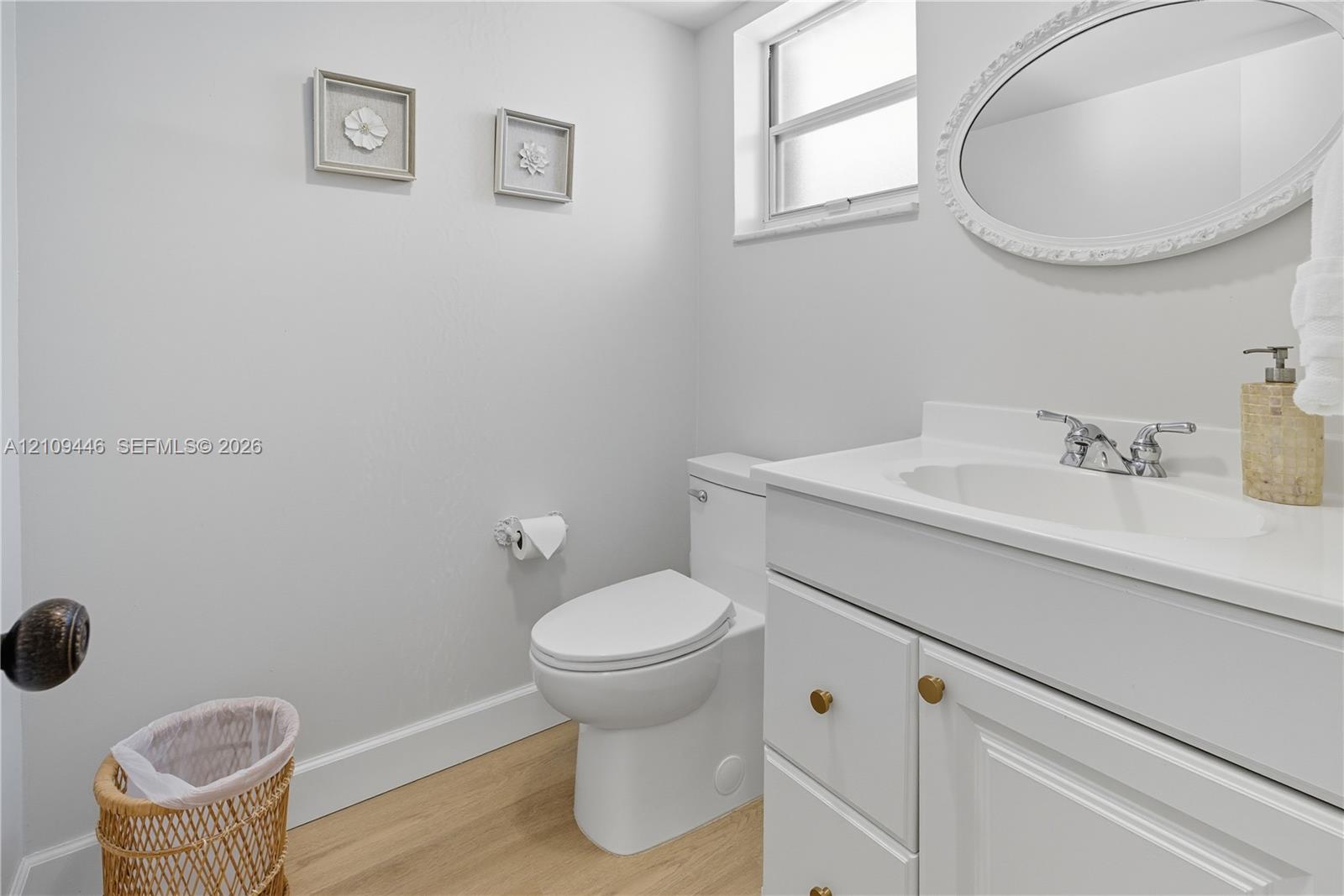 White vanity with integrated sink, oval wall-mounted mirror, wood-finish flooring, and single-pane window