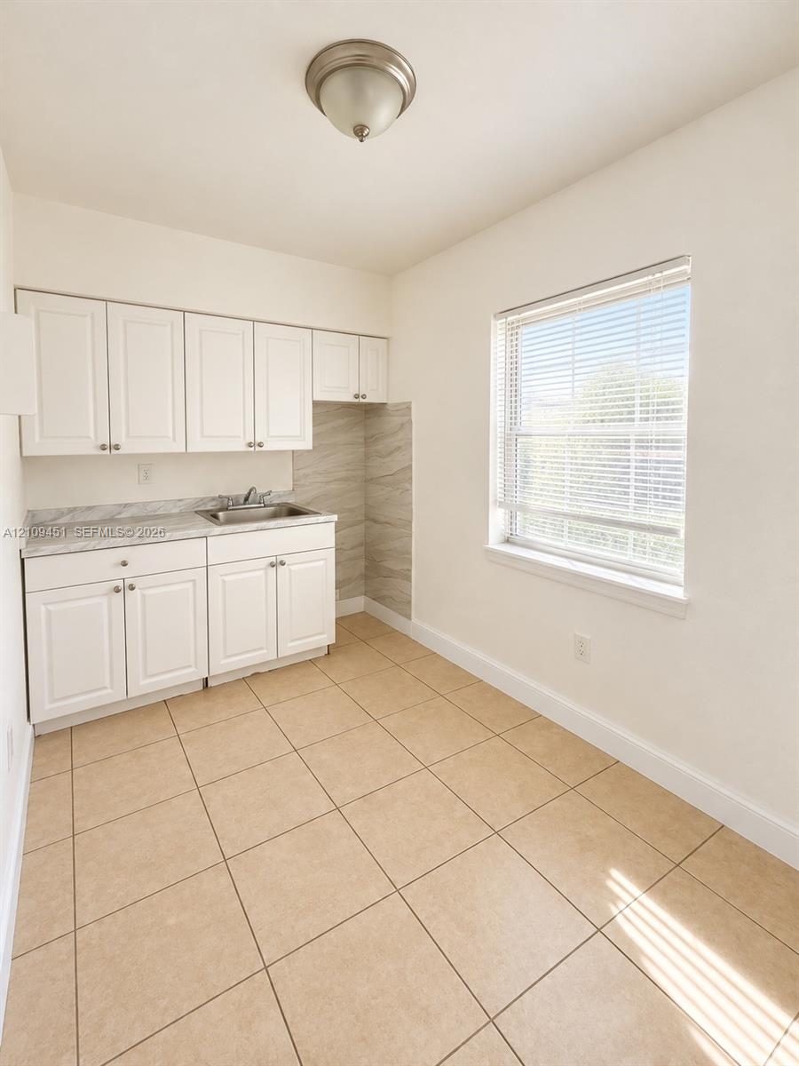 White shaker-style cabinetry, neutral stone-patterned countertops, stainless steel sink, large-format ceramic tile flooring, and a flush-mount ceiling light fixture