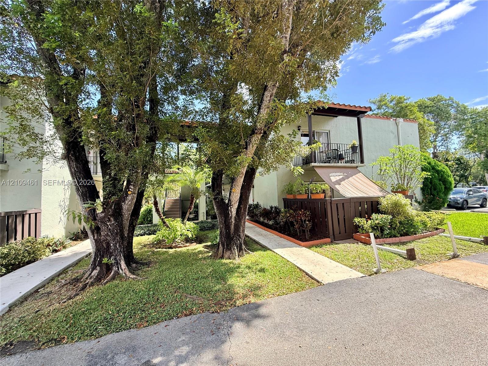 Two-story exterior featuring light-colored stucco siding, a tiled roofline, second-level balcony with railings, ground-level patio with privacy fencing, and mature trees