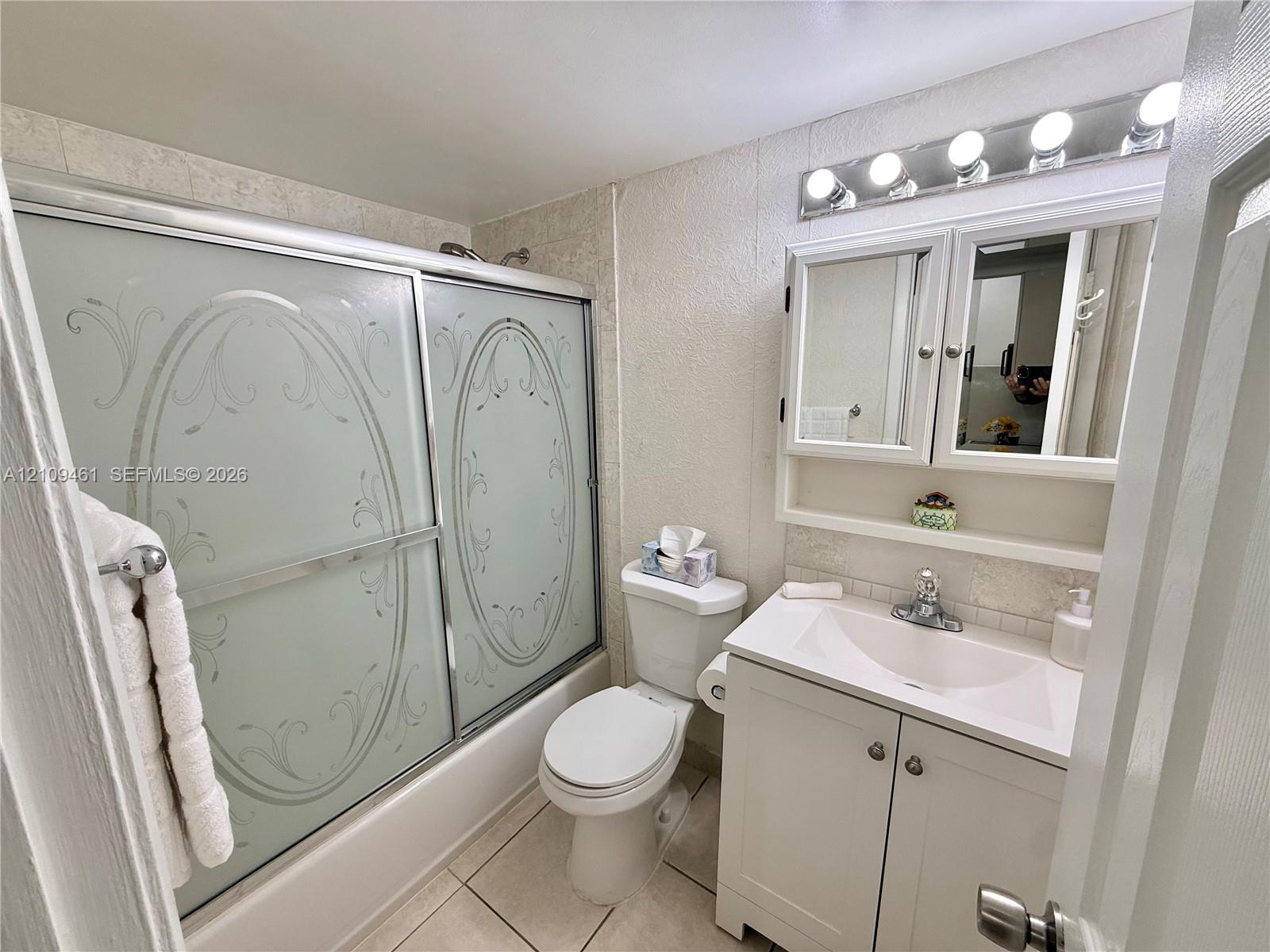 Bathroom featuring a tub-shower combination with etched glass sliding doors, a white vanity cabinet with an integrated sink, a multi-bulb vanity light fixture, and tile flooring