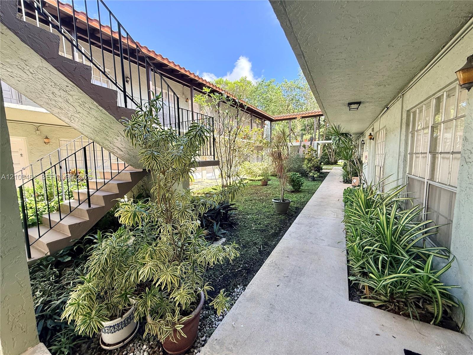 Covered exterior walkway with a concrete path, exterior staircase featuring metal railings, and landscaped courtyard area