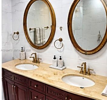 Double vanity with stone countertop, dual undermount sinks, brass-toned fixtures, wood cabinetry, and twin oval mirrors with ornate gold-tone frames