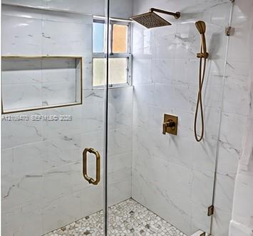 Walk-in shower featuring white large-format tile walls, mosaic stone tile flooring, a built-in shower niche, frameless glass enclosure, and metallic gold-finish fixtures including a rainfall showerhead and handheld sprayer